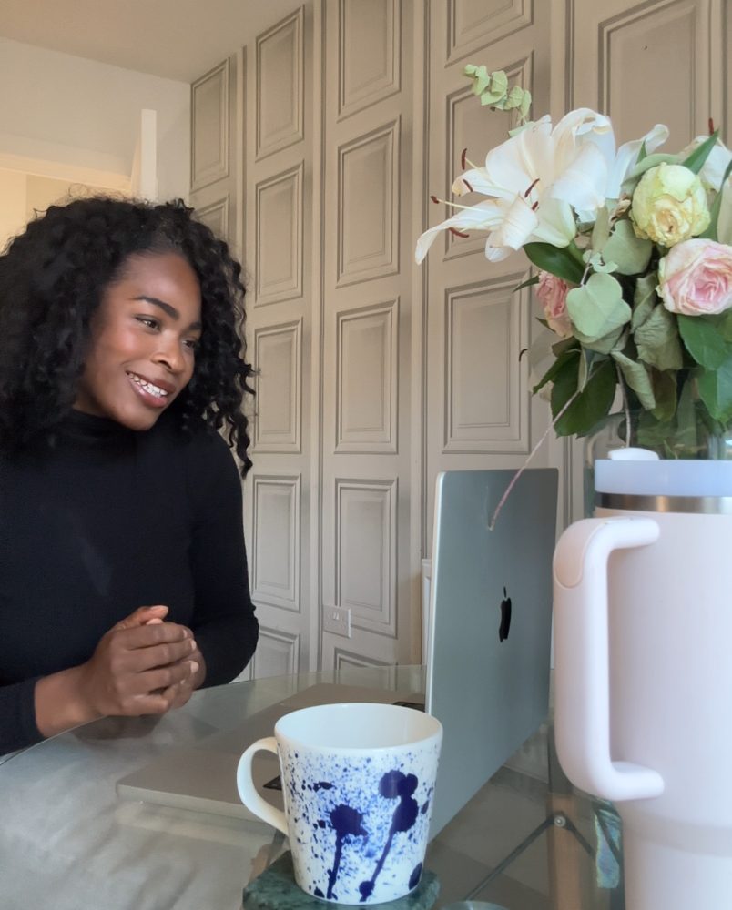 Jade smiling on a video call at her glass dining table, with fresh flowers and a laptop